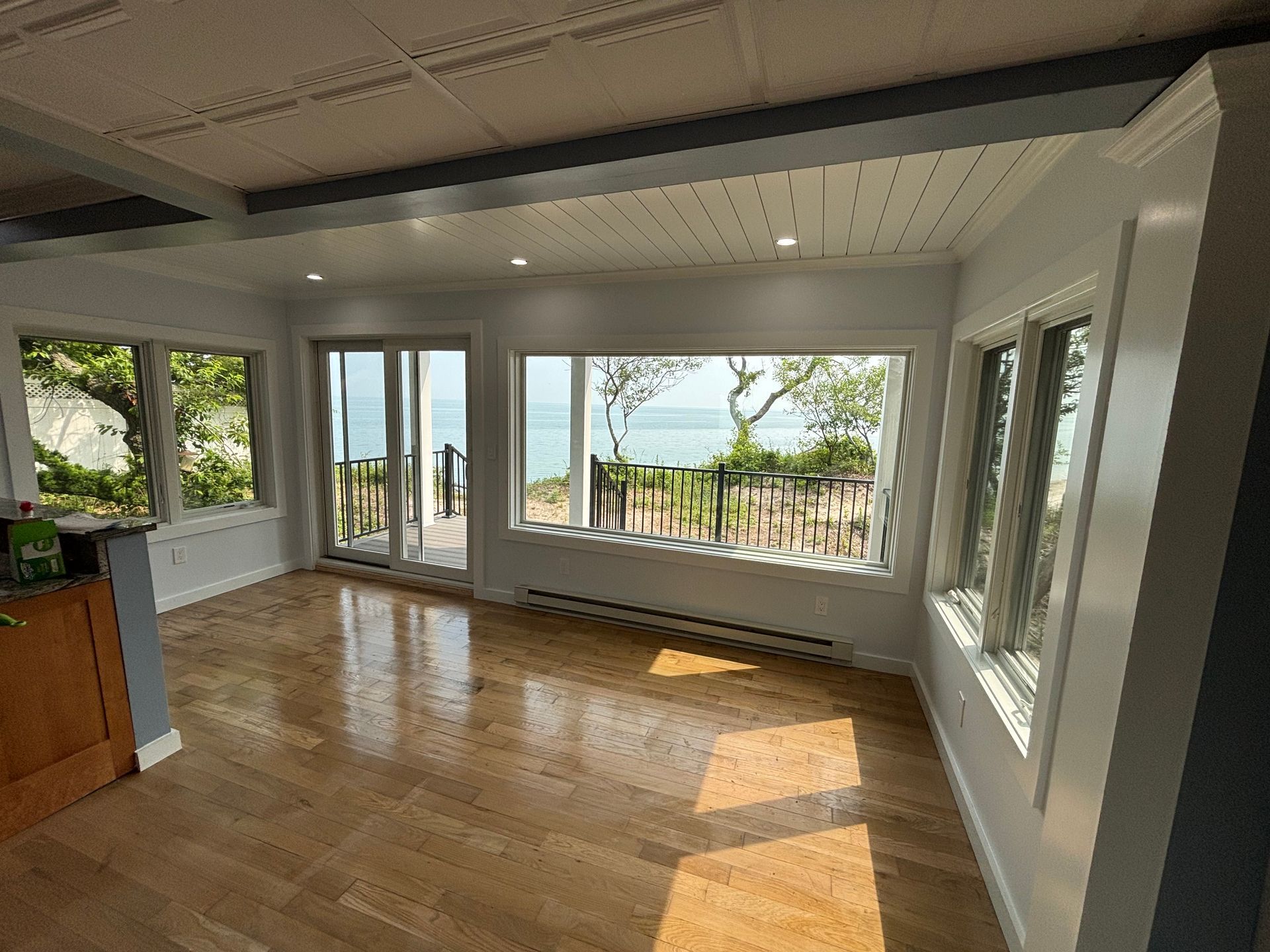 Bright empty sunlit room with hardwood floors and large windows overlooking a balcony and trees.
