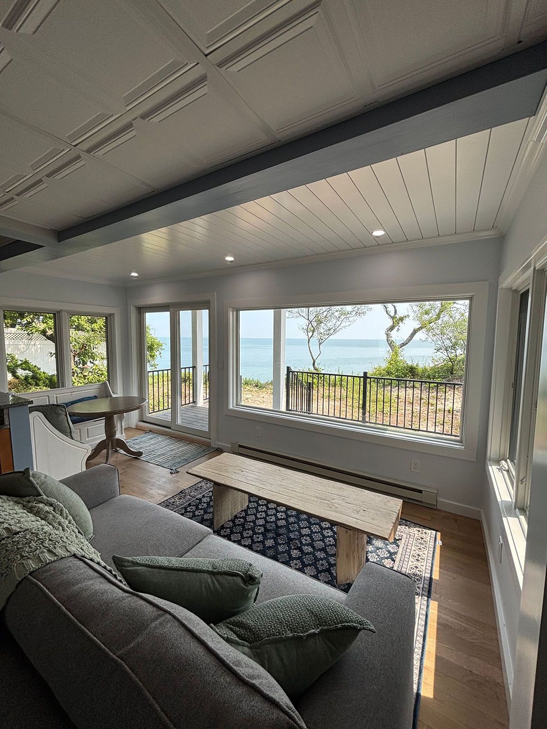 Modern living room with gray sectional, wooden coffee table, and large windows overlooking water and greenery.