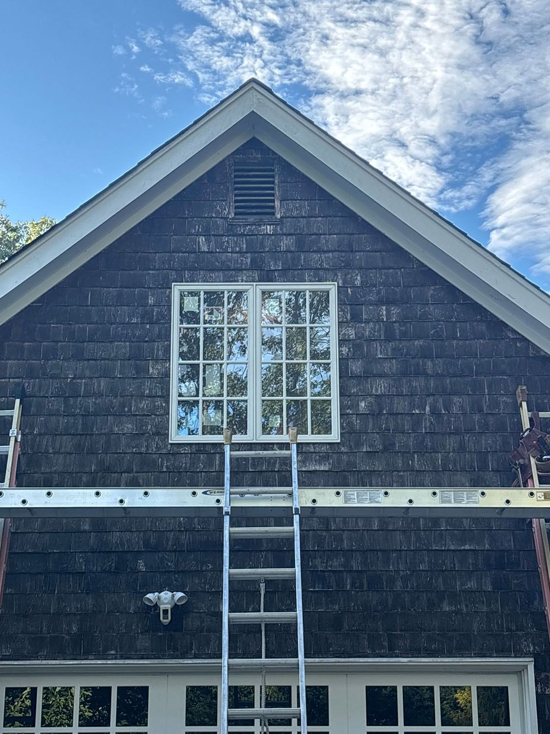 Ladder leaning against dark-shingled house under gabled roof, with a wide window and blue sky above.