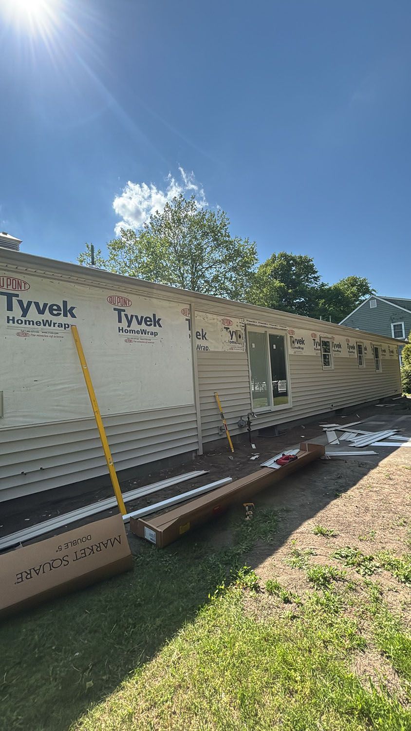 House under construction with Tyvek wrap, windows installed, and lumber on a sunny day.