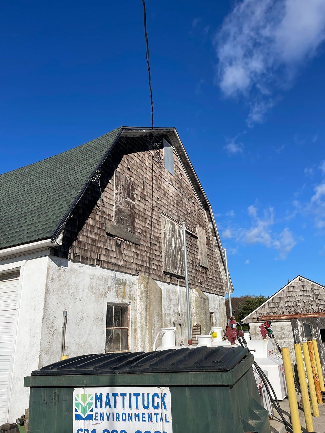 Weathered building with white lower walls and dark shingled roof under a blue sky, with a dumpster in front.