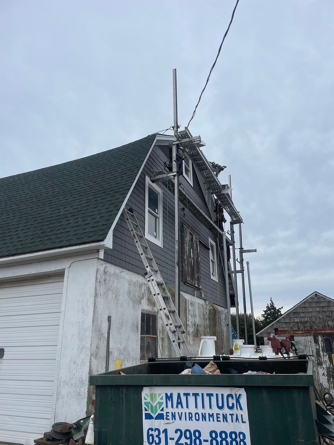 Damaged house roof with exposed framing and debris beside a blue dumpster under repair.