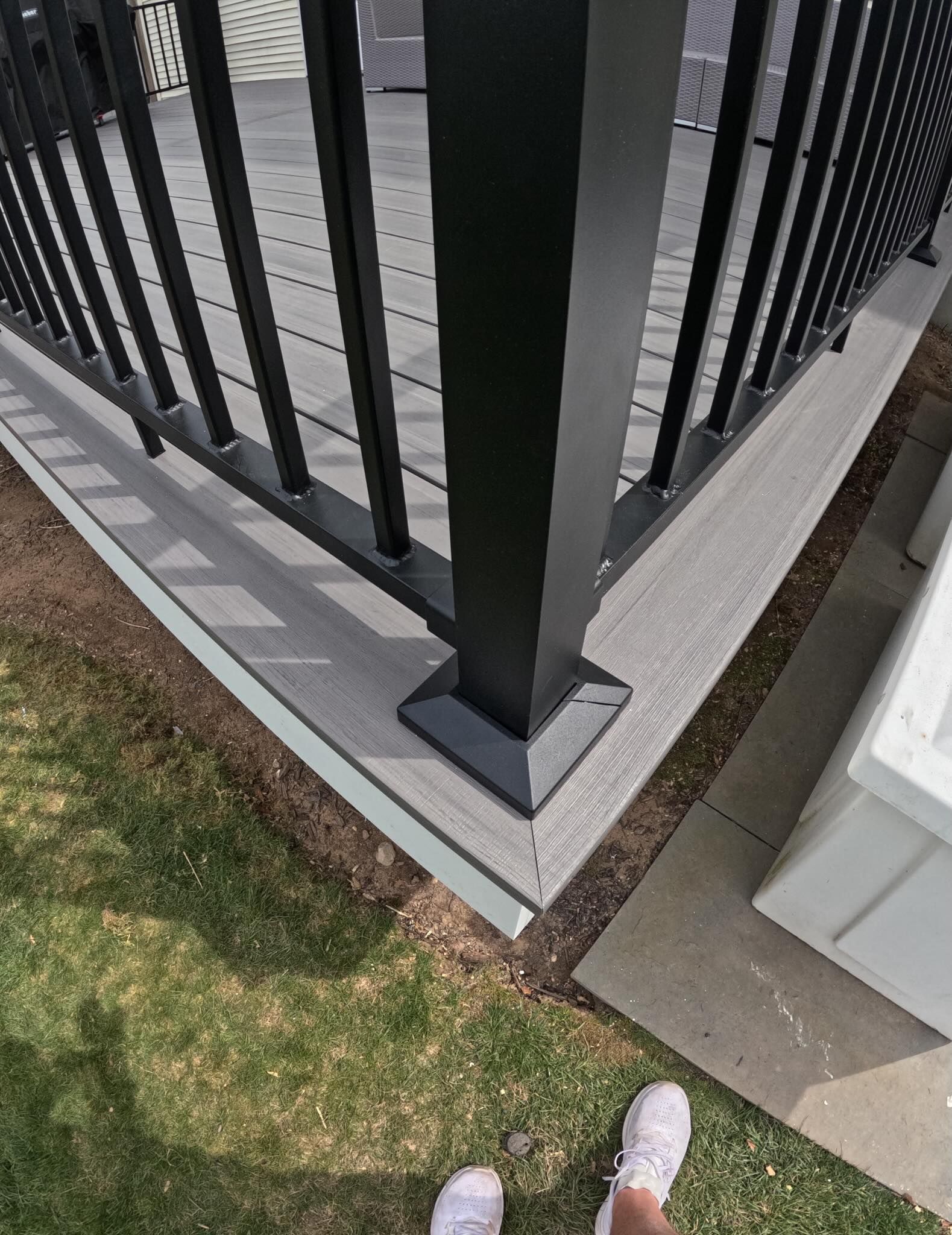 Corner of a black metal balcony railing above a paved patio and grass, with pink shoes visible below