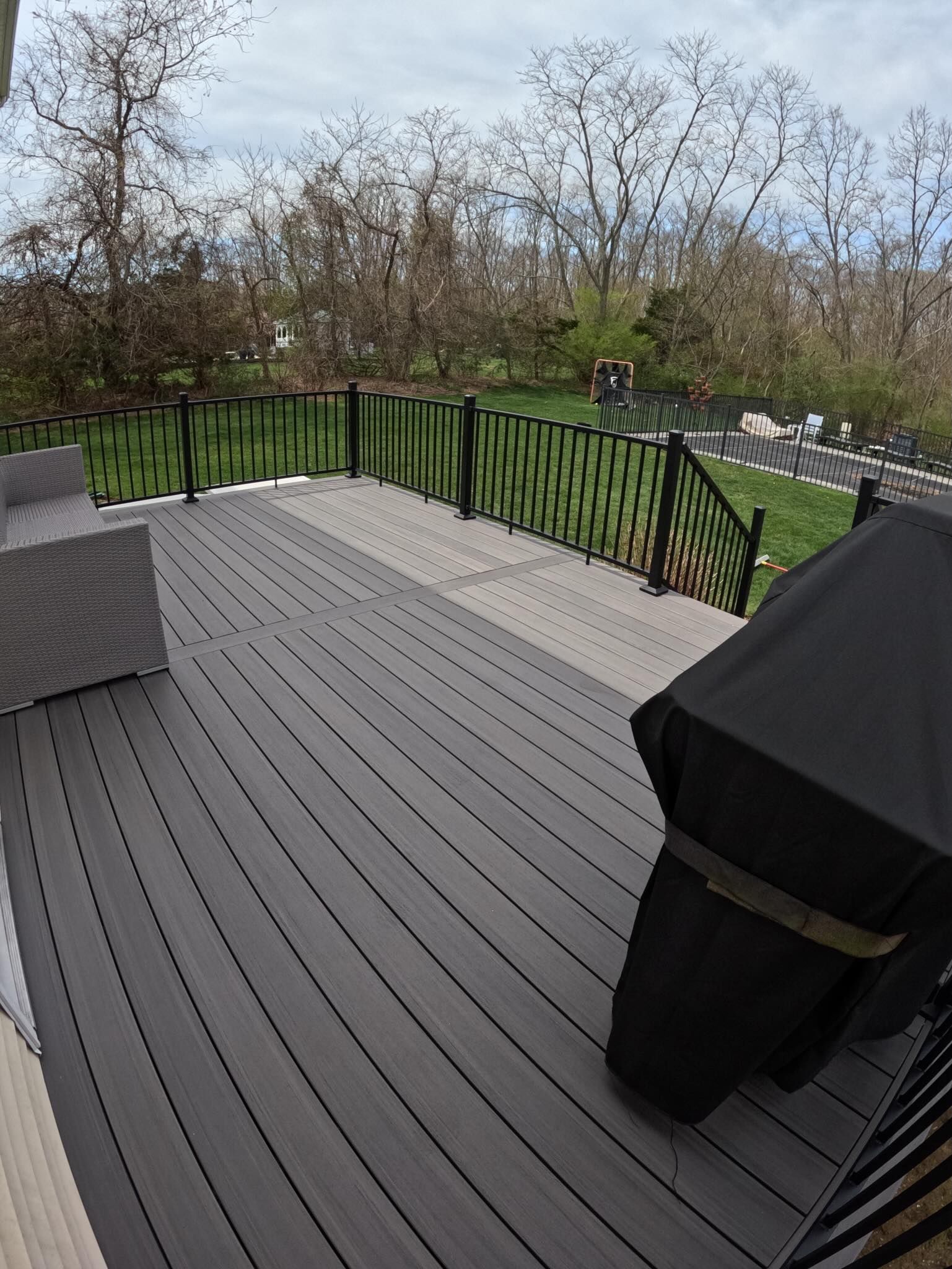 Wooden deck with black railing, covered grill, and wooded yard beyond.