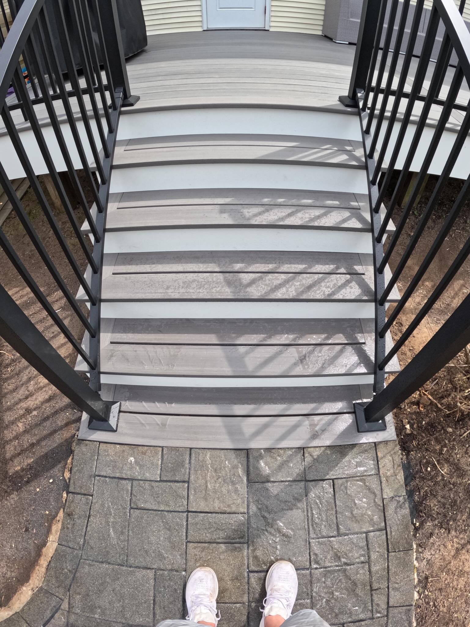 Outdoor staircase and stone walkway viewed from above, with black railings and a person's white shoes at the bottom.