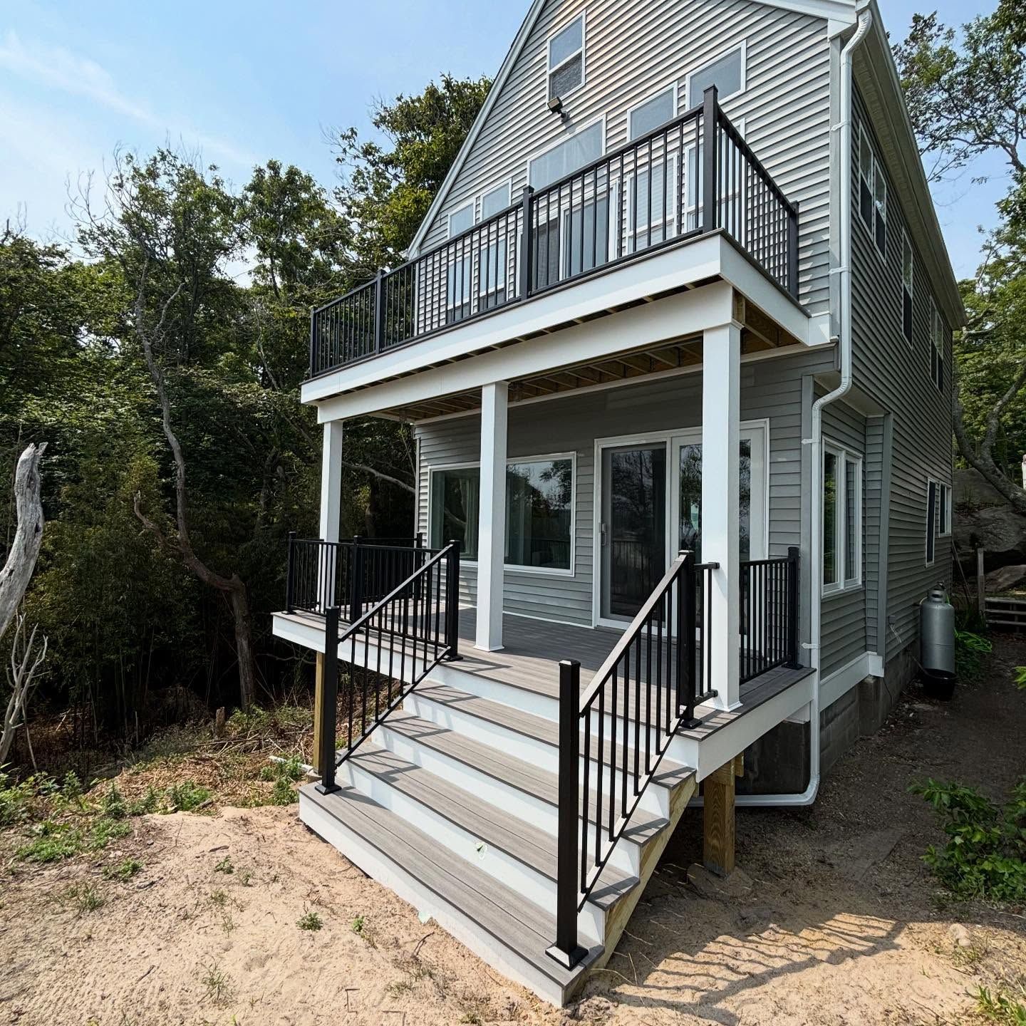 Two-story gray house with front porch, stairs, and upper balcony surrounded by trees