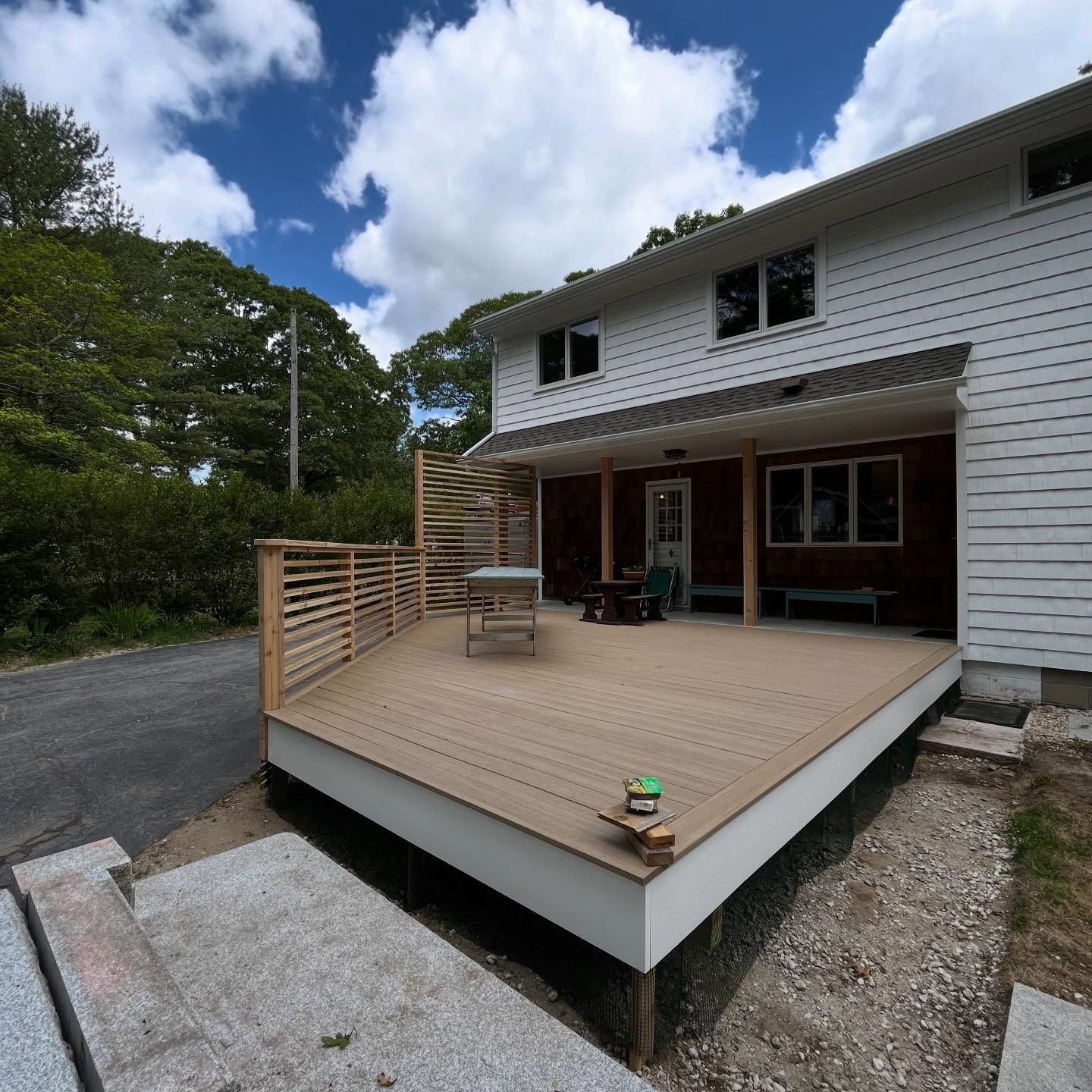 Wooden backyard deck beside a white house, with railing, gravel, and trees under a cloudy sky