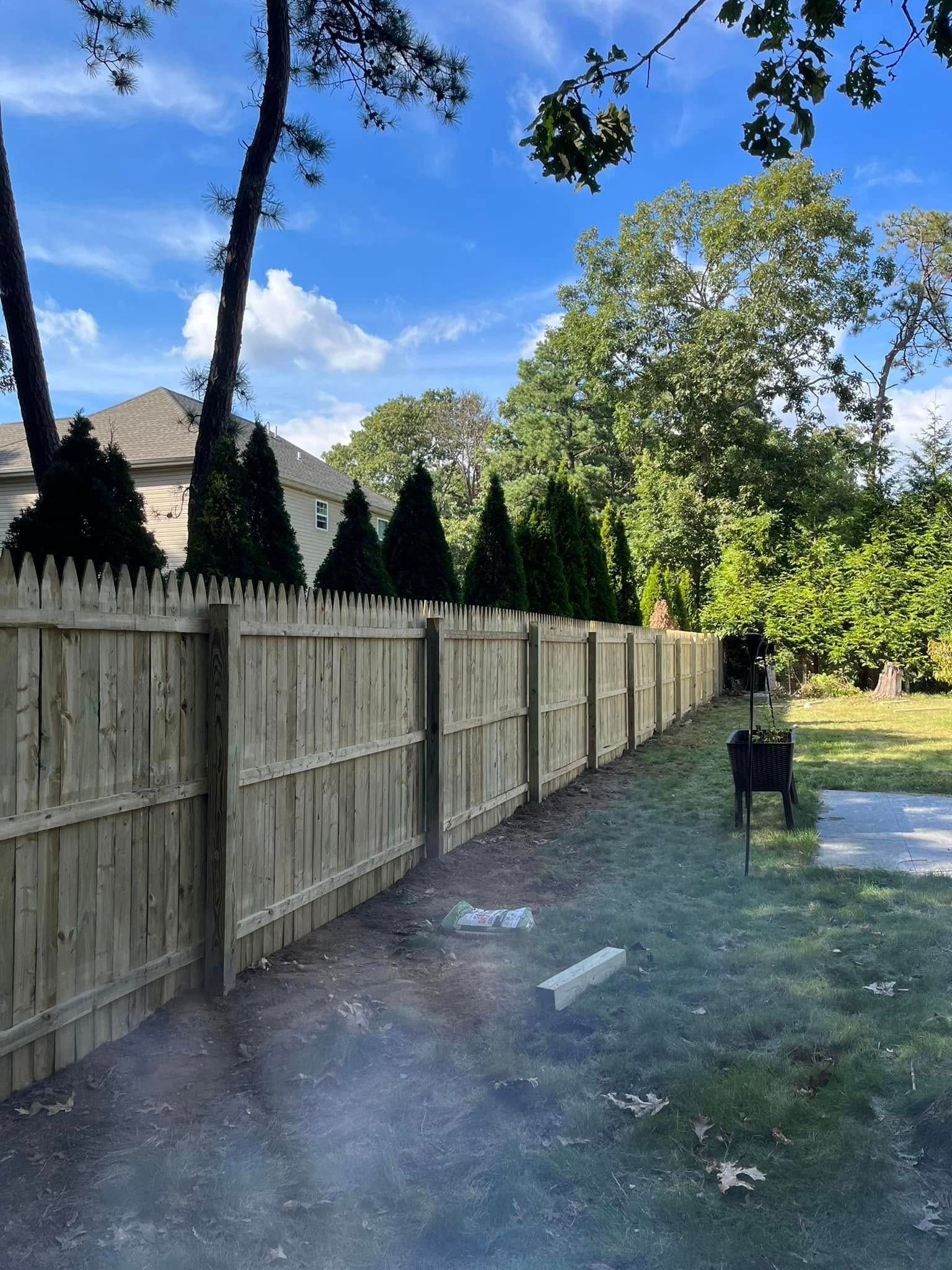 Wooden privacy fence beside a grassy yard with trees and a blue sky on a sunny day