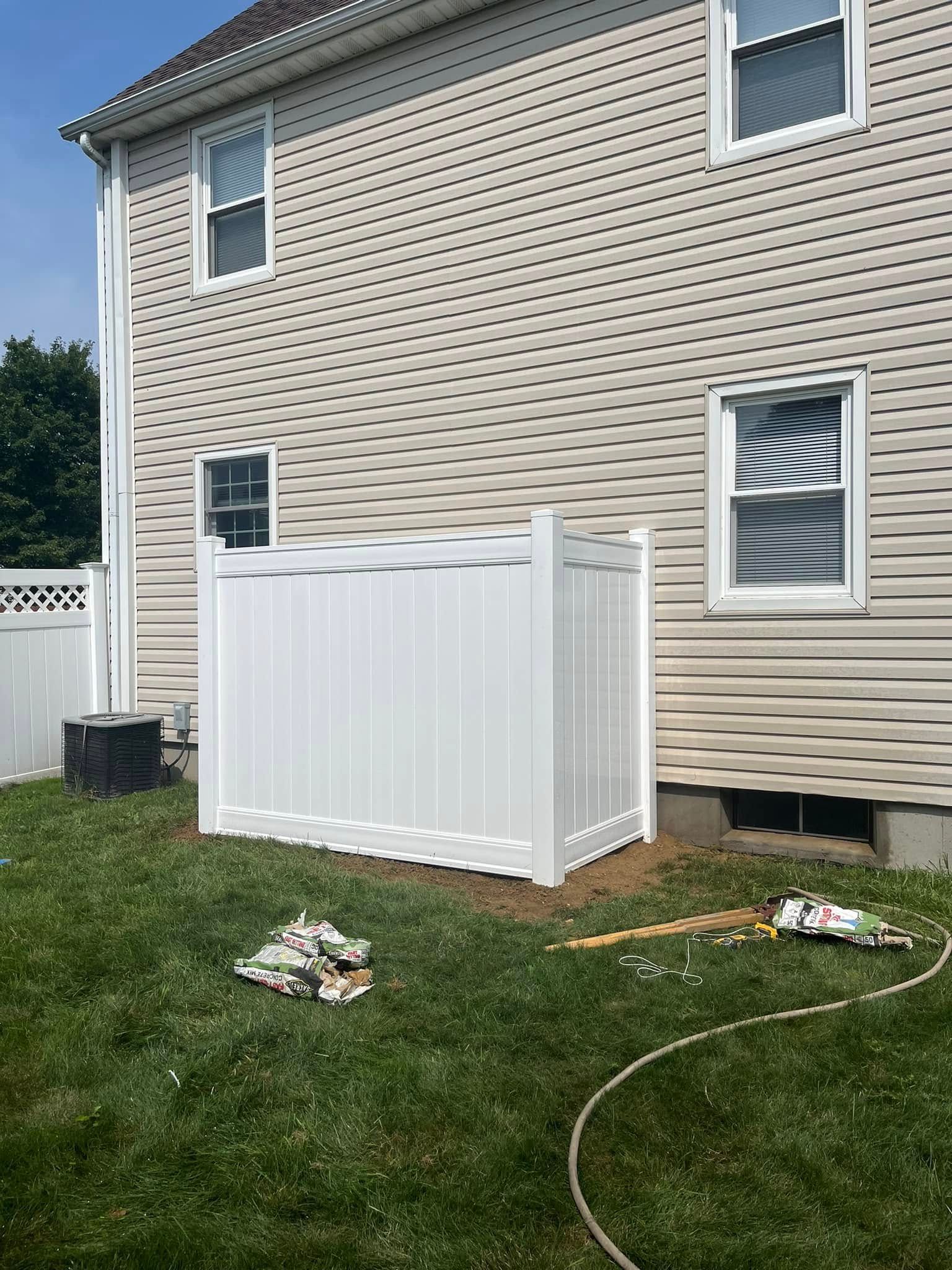 White outdoor storage enclosure against a beige house beside a grassy yard and white fence.