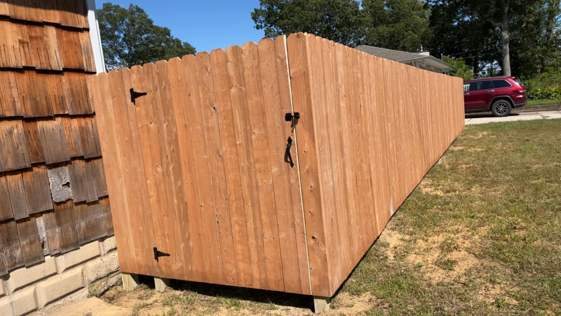 Wooden fenced enclosure with a gate in a grassy yard, red car parked in the background.