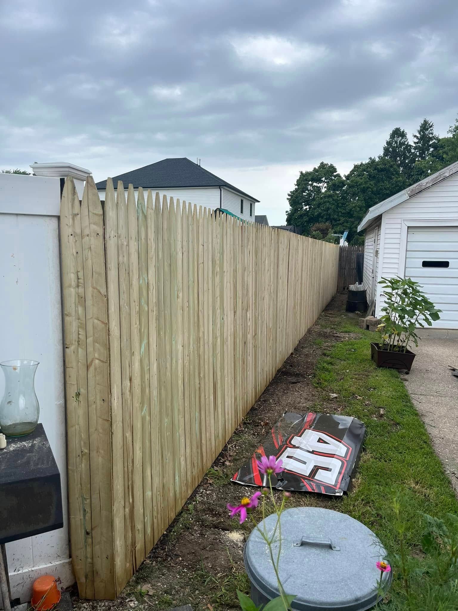 Backyard with new wooden fence beside a house, grass strip, and utility covers in the foreground