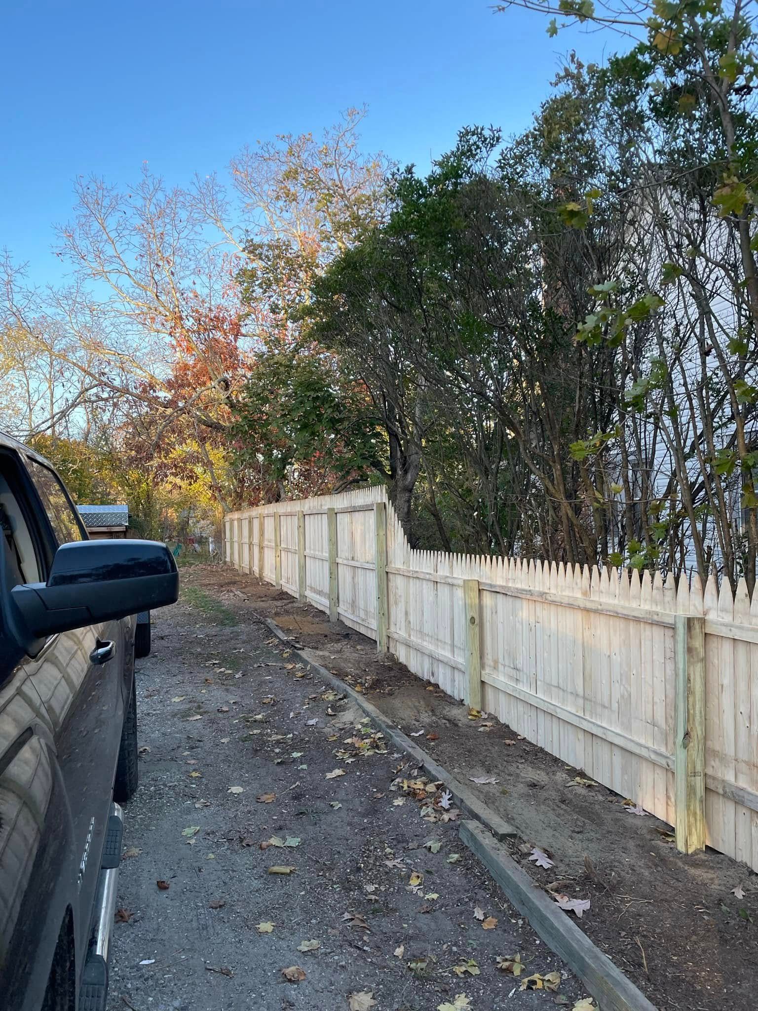 White fence along a dirt roadside beside a parked truck, with autumn trees overhead.