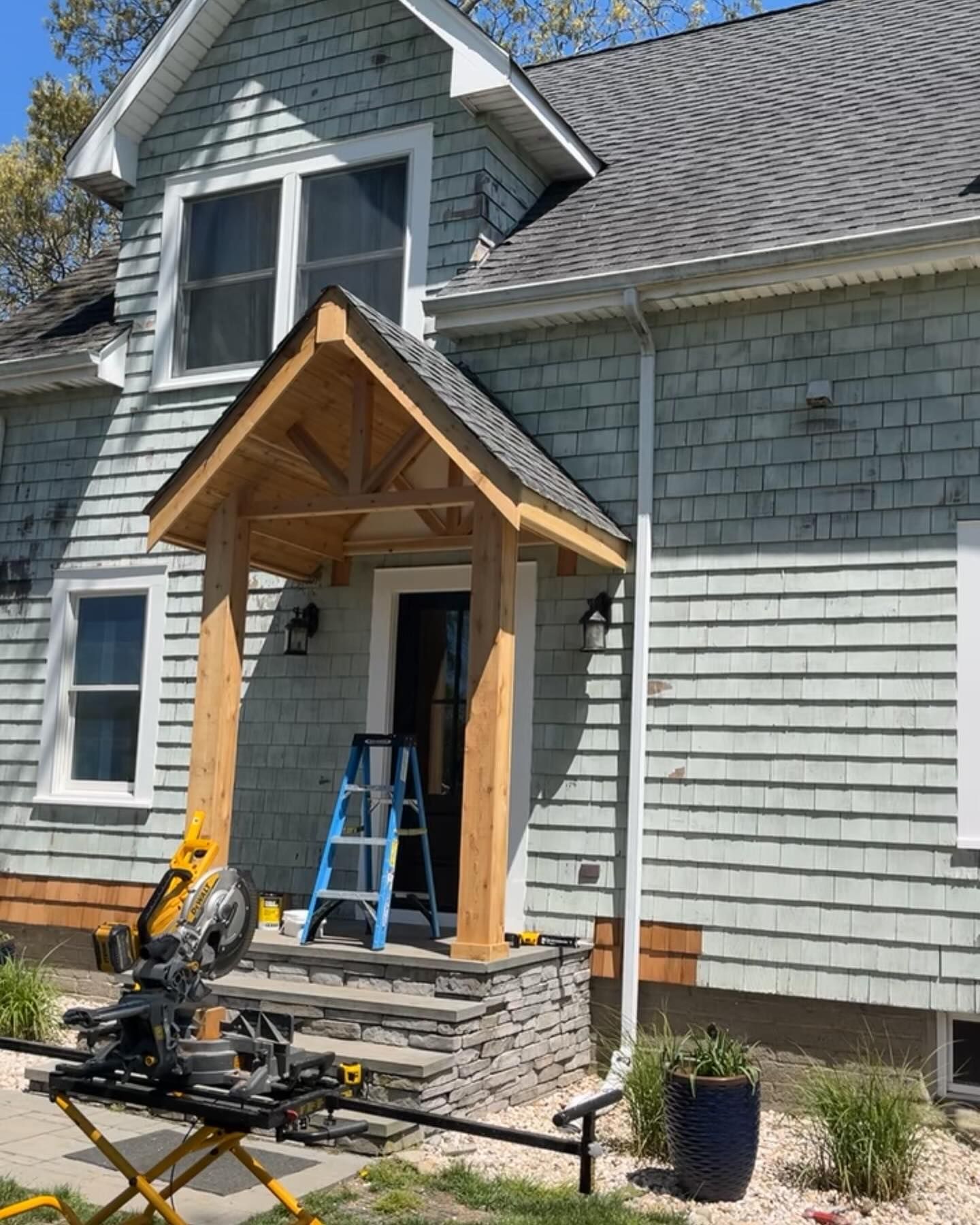 Front porch under construction with wooden entry canopy, ladder, and power tools beside a gray house