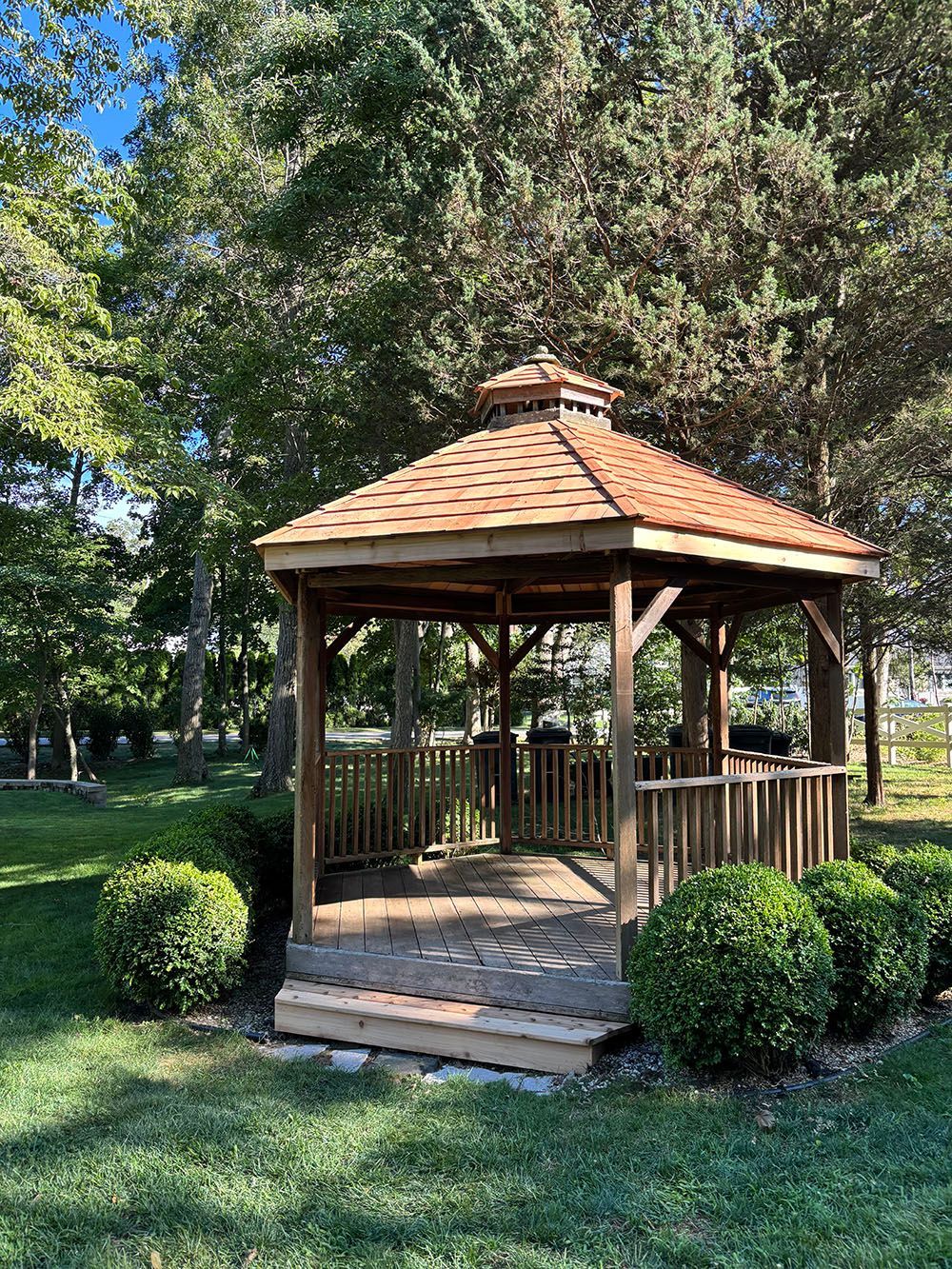 Wooden gazebo with a shingled roof in a green park, surrounded by trees and shrubs.