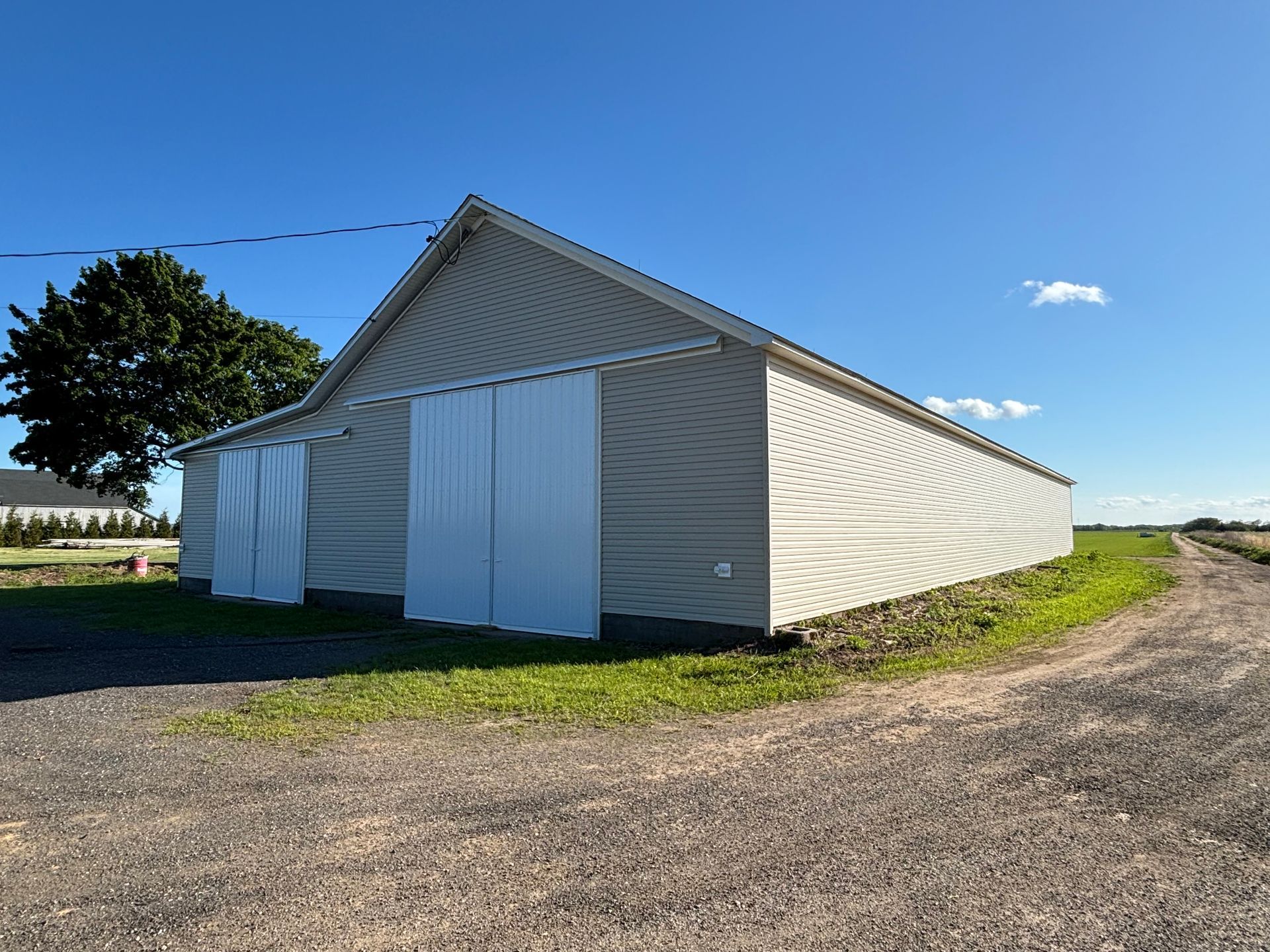 White barn on a gravel road under a clear blue sky, with a tree and open field nearby.