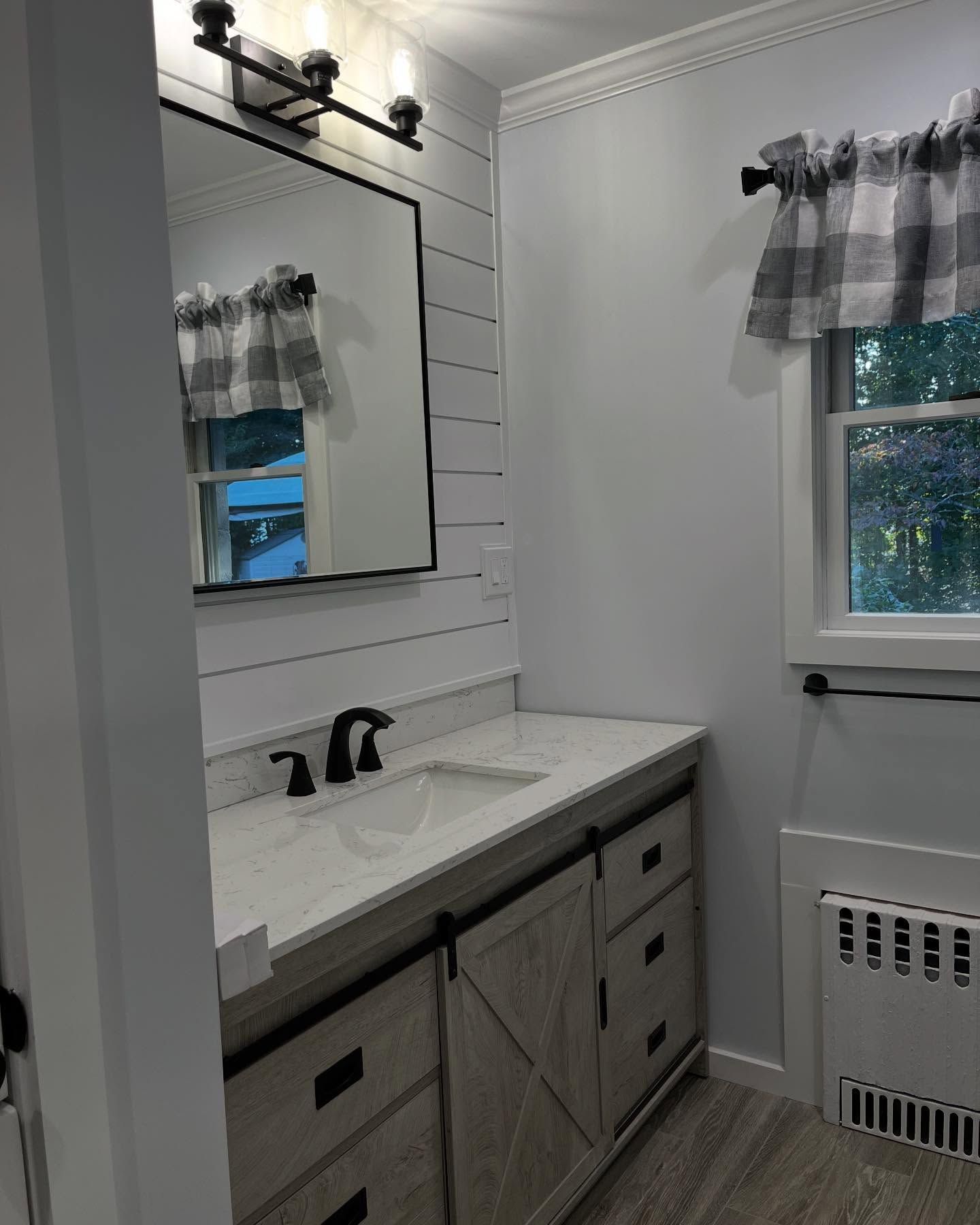 Modern bathroom with vanity, black sconces, shiplap wall, mirror, and window with sheer curtains.