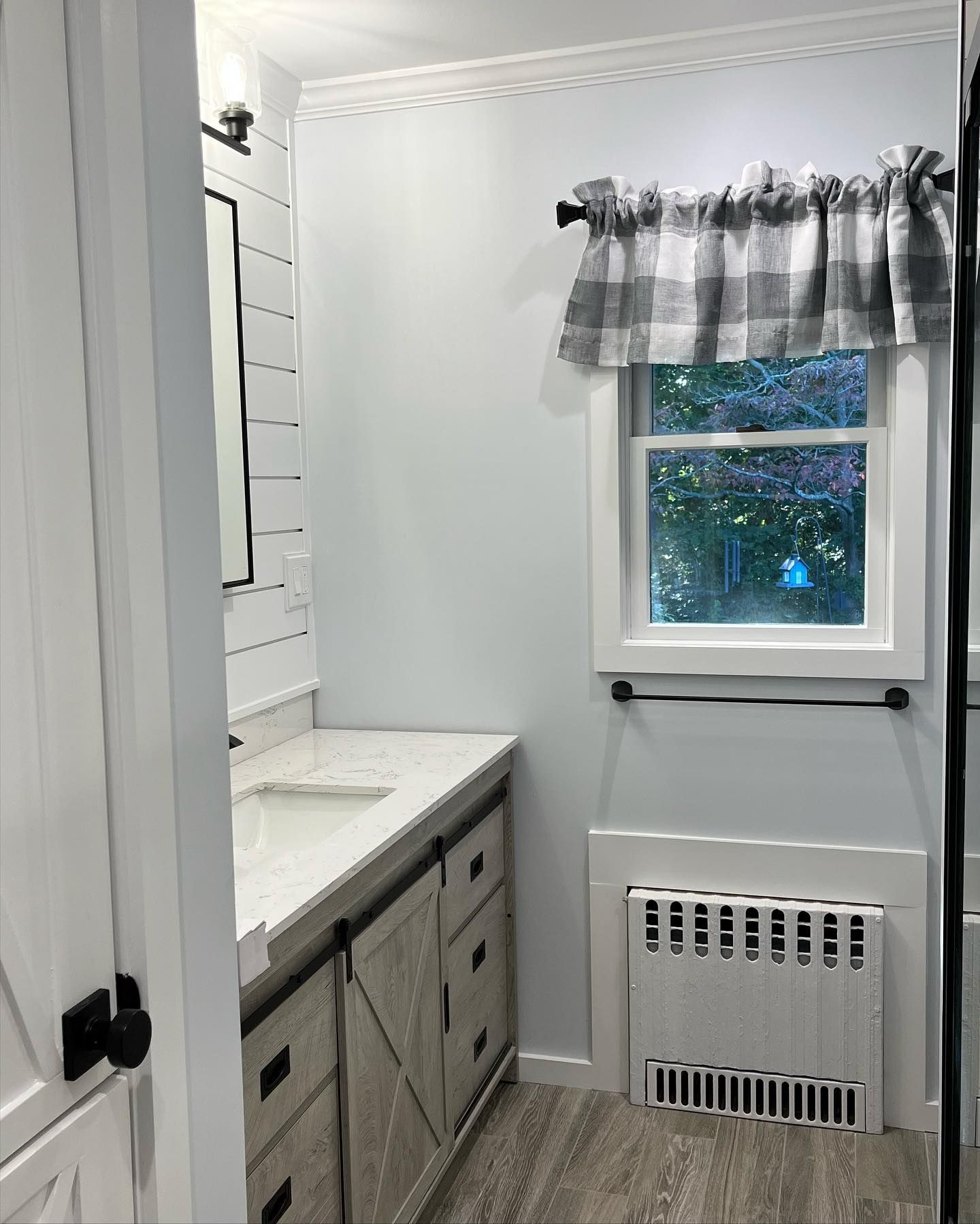 Small bathroom with gray vanity, white walls, window curtain, and wall heater beneath the window.