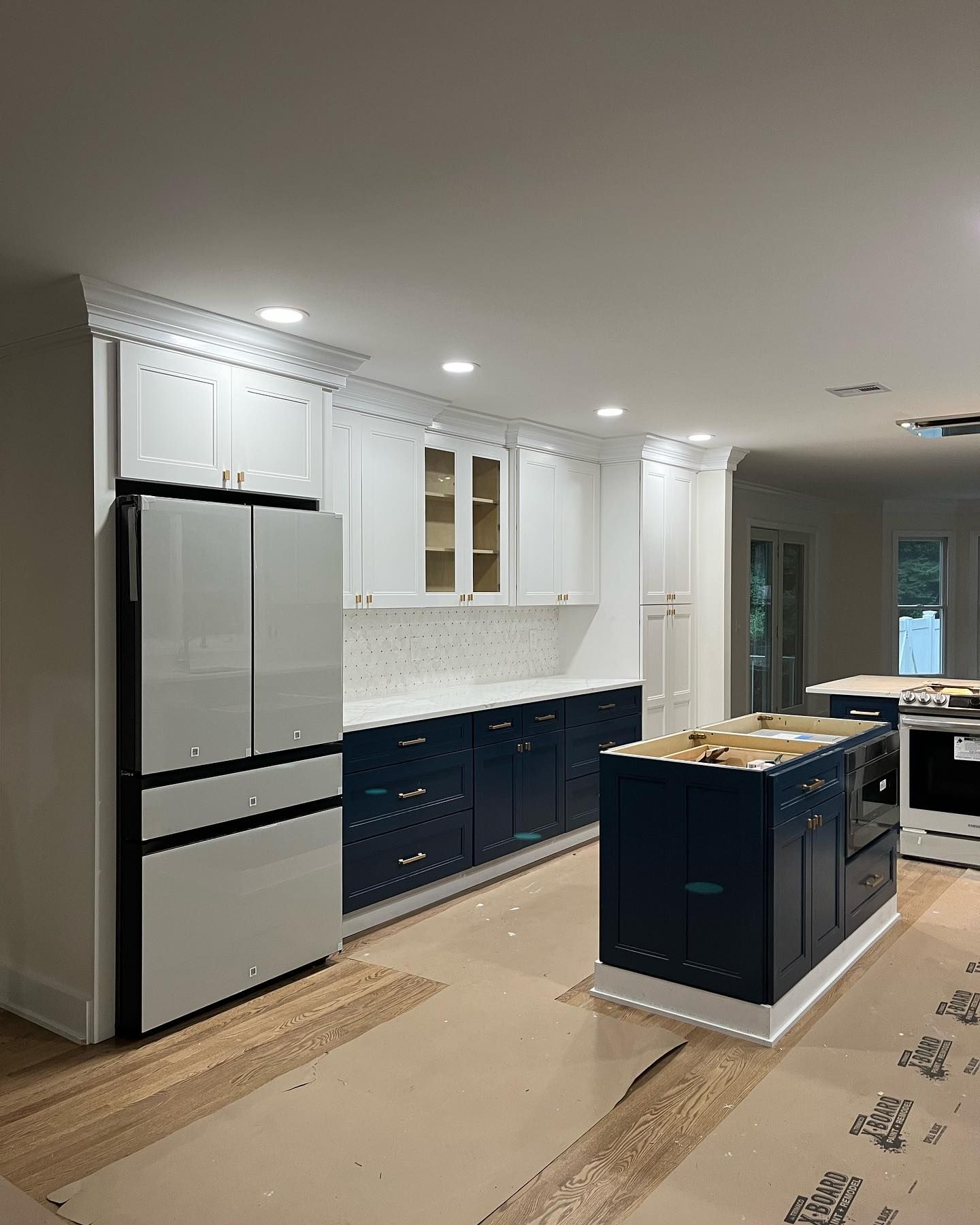 Modern kitchen with white cabinets, navy island, stainless fridge, and protective floor covering during installation