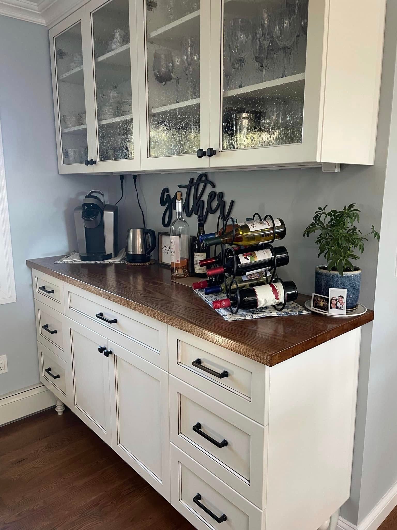 White kitchen buffet with brown countertop, glass-front cabinets, coffee setup, and a potted plant.
