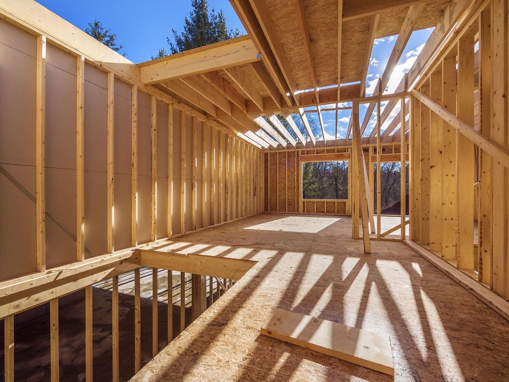 Interior of a wooden house under construction, with exposed framing and sunlight casting shadows through the roof.