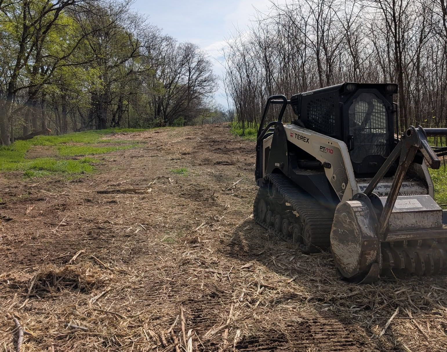 Compact track loader clearing a dirt path through a wooded area