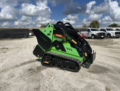 A red Toro tracked stand-on skid steer loader with an open grapple attachment, parked on a grassy lot near a building.