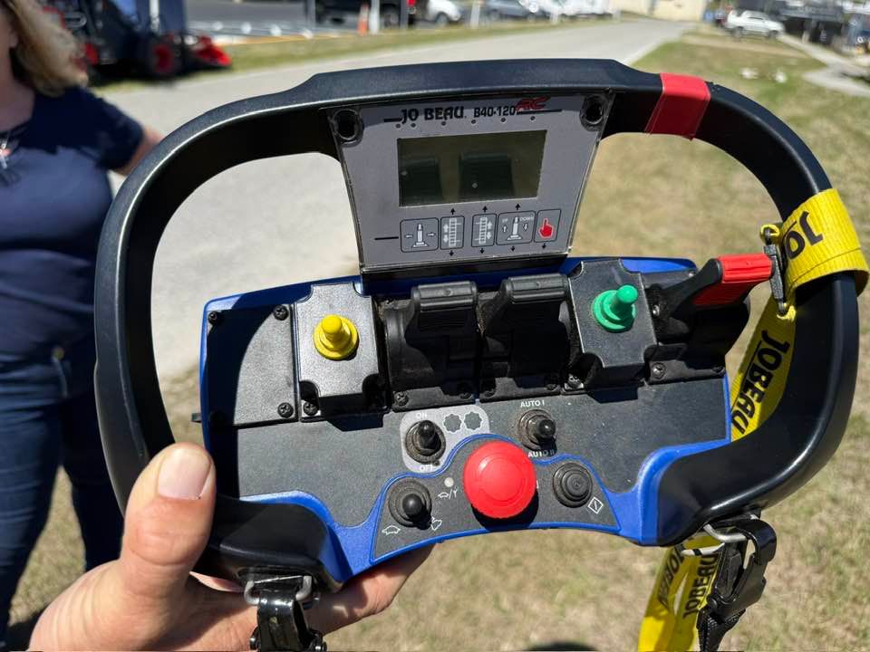 A person holds a remote control unit with a display screen, joysticks, and a prominent red emergency stop button.