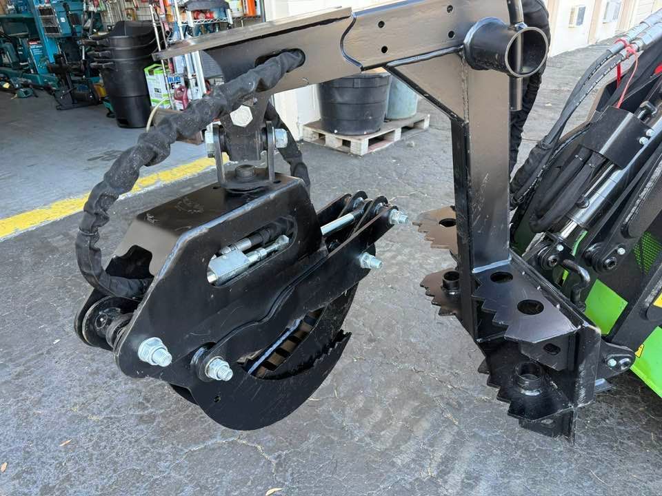 A black hydraulic grapple attachment mounted on a tractor, positioned over a paved workshop floor.