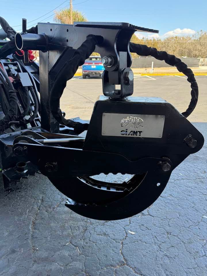 A black hydraulic grapple attachment mounted on machinery, centered in an outdoor parking lot setting.