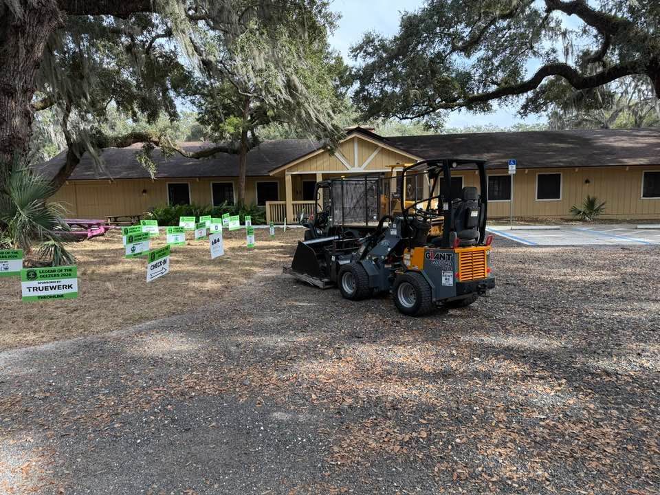 A yellow skid steer loader sits on a gravel lot in front of a tan, single-story building with many lawn signs nearby.