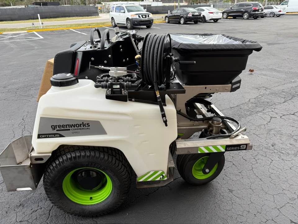 A side view of a white Greenworks commercial spreader-sprayer machine parked on an asphalt lot.