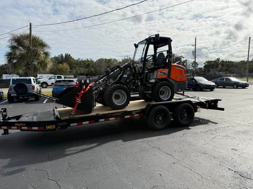 An orange and black articulated loader secured with red straps on a flatbed trailer in a parking lot.