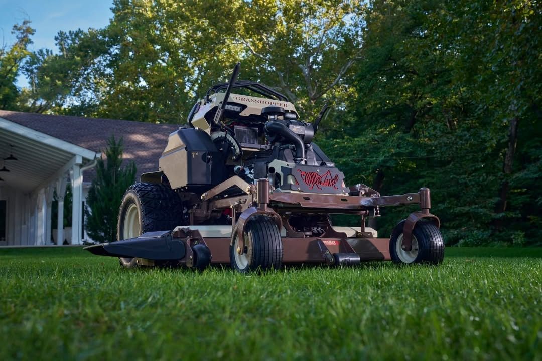 A brown commercial zero-turn mower sits on a green lawn in front of a house and trees.