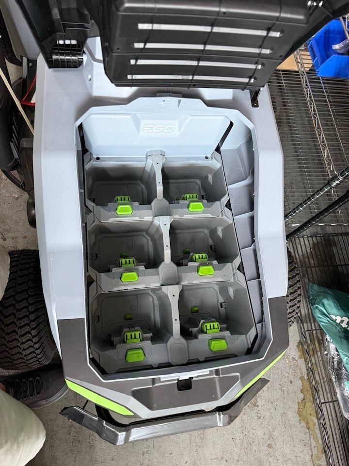An open battery compartment of a lawn mower with six empty gray slots featuring lime green locking tabs.