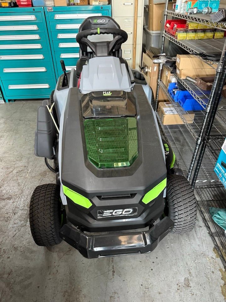 An EGO electric riding lawn mower parked indoors in a workshop or garage, with teal tool cabinets and shelving nearby.