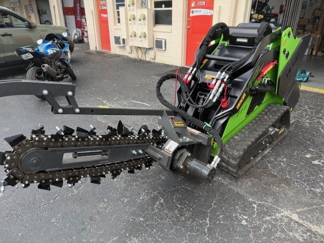 A lime green compact track loader equipped with a trencher attachment is parked on an outdoor pavement.
