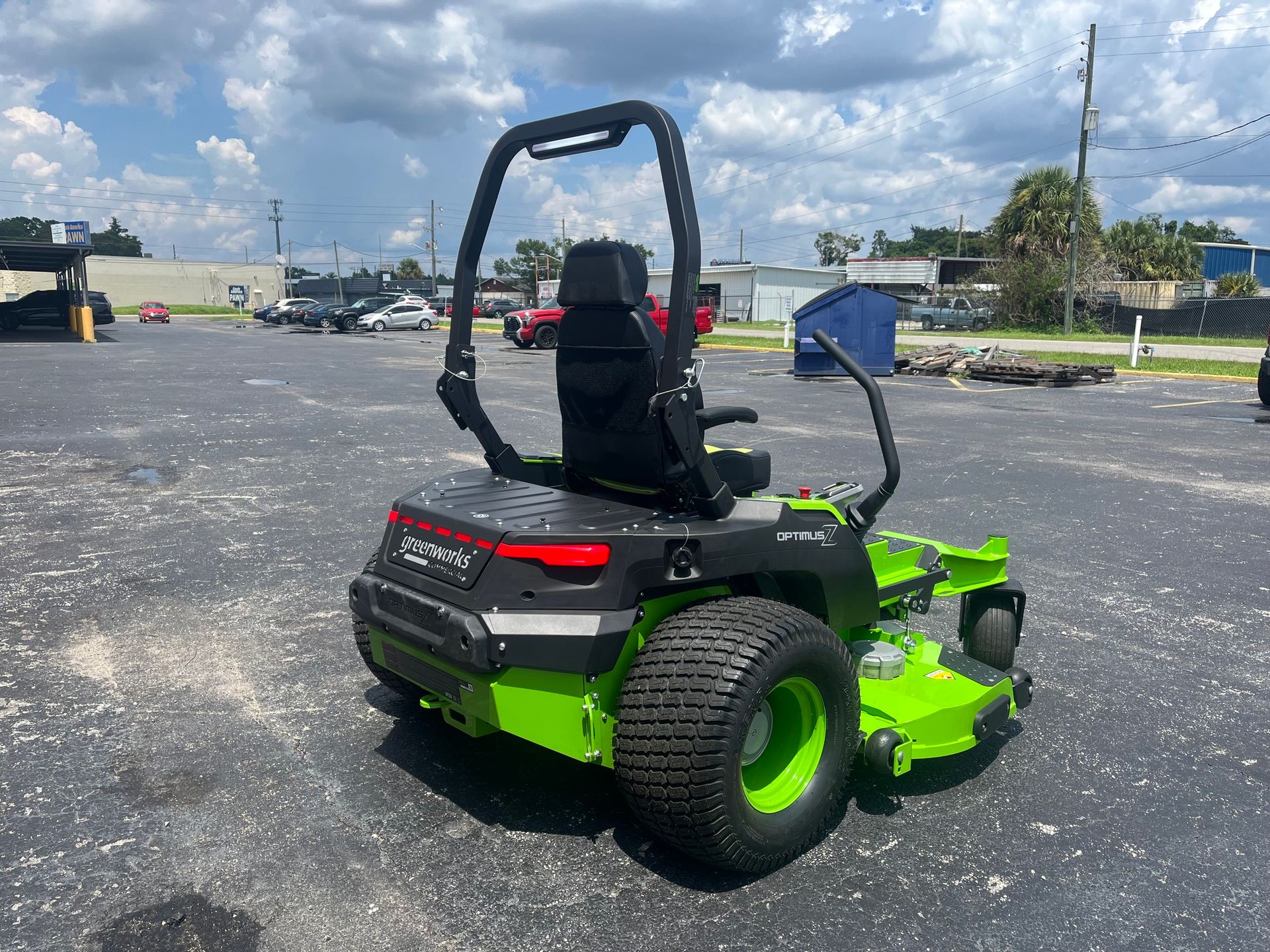 A lime-green and black zero-turn lawn mower parked on an asphalt lot under a blue sky with clouds.
