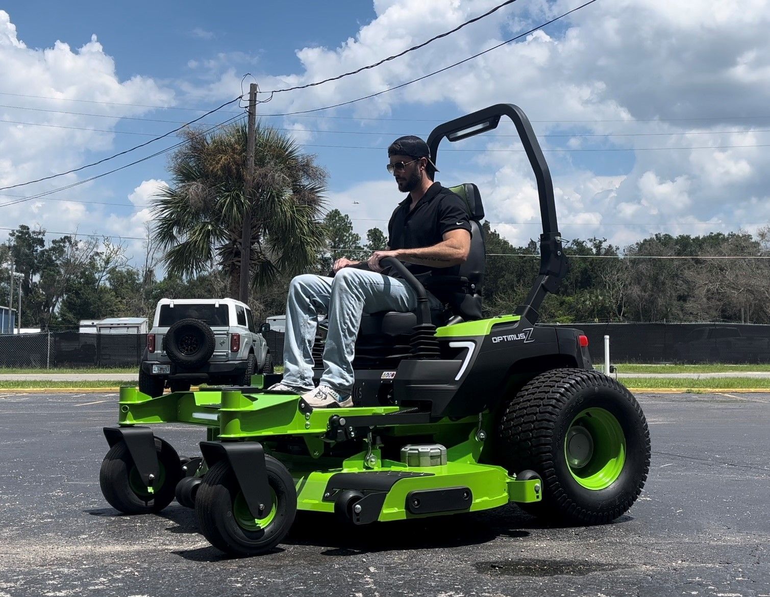 A person wearing a black shirt and jeans drives a bright lime green zero-turn lawn mower in a paved lot.