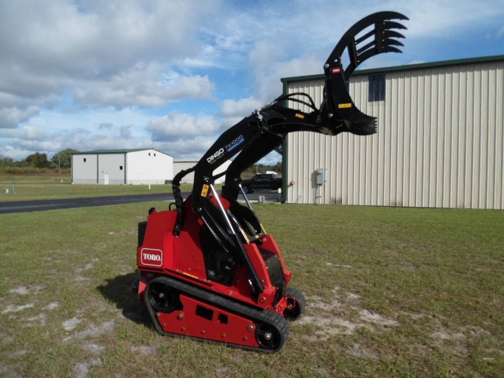 A red Toro compact utility loader with a raised grapple attachment parked on a grassy field near metal buildings.