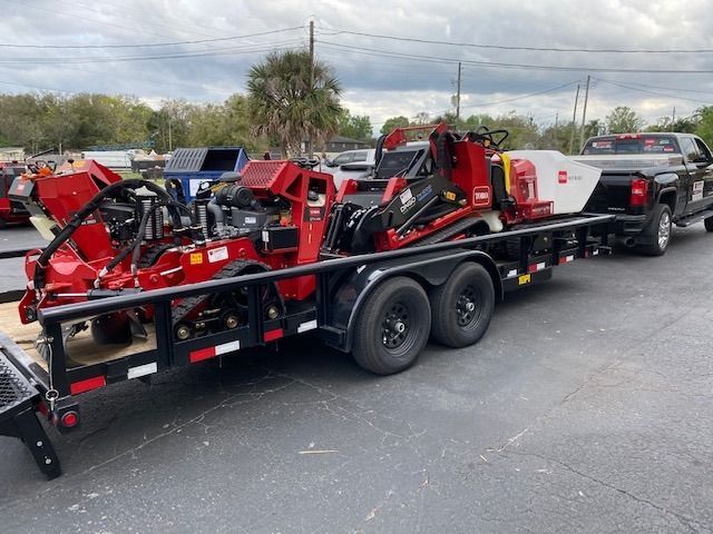 A red industrial trenching machine loaded on a flatbed trailer attached to a pickup truck parked in an outdoor lot.