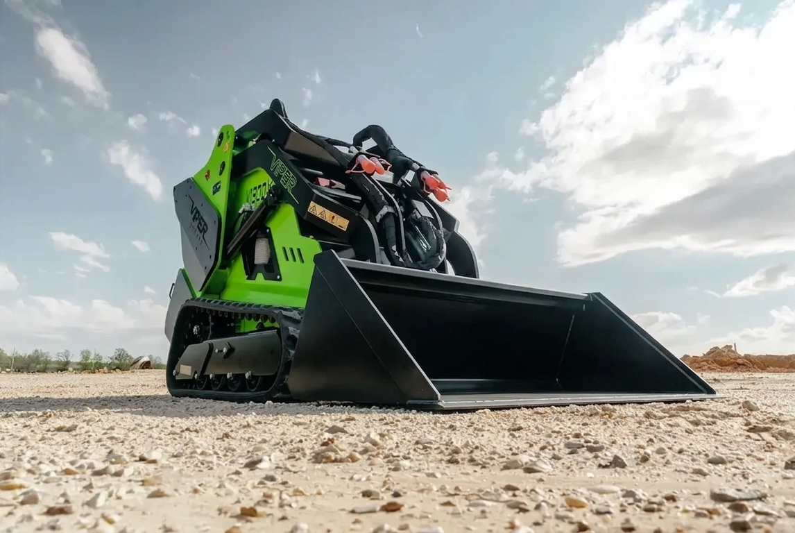 Black-and-green tracked skid-steer loader with front bucket on rocky ground under a cloudy sky