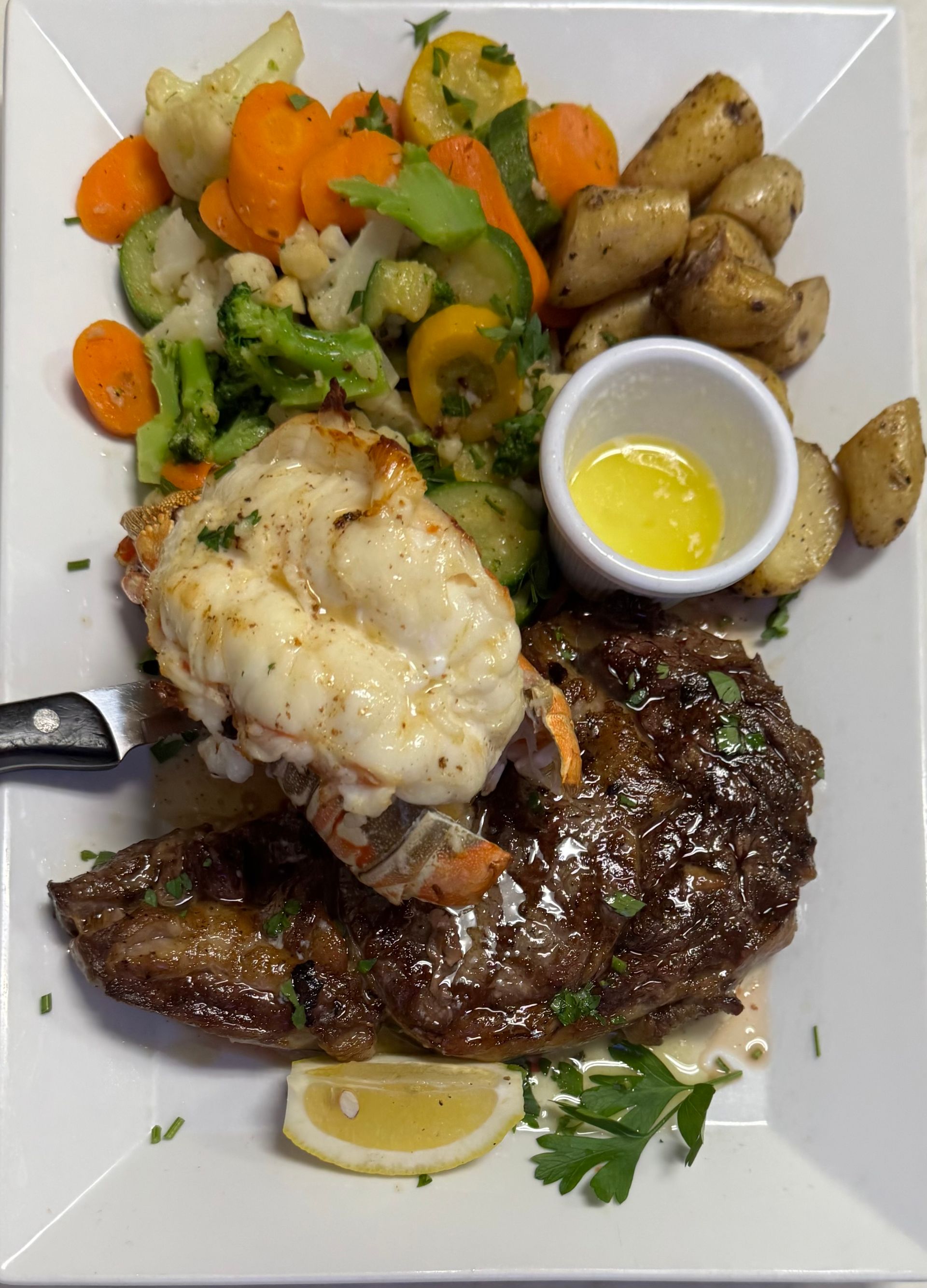 Steak and lobster surf and turf with vegetables, potatoes, and melted butter on a white plate.