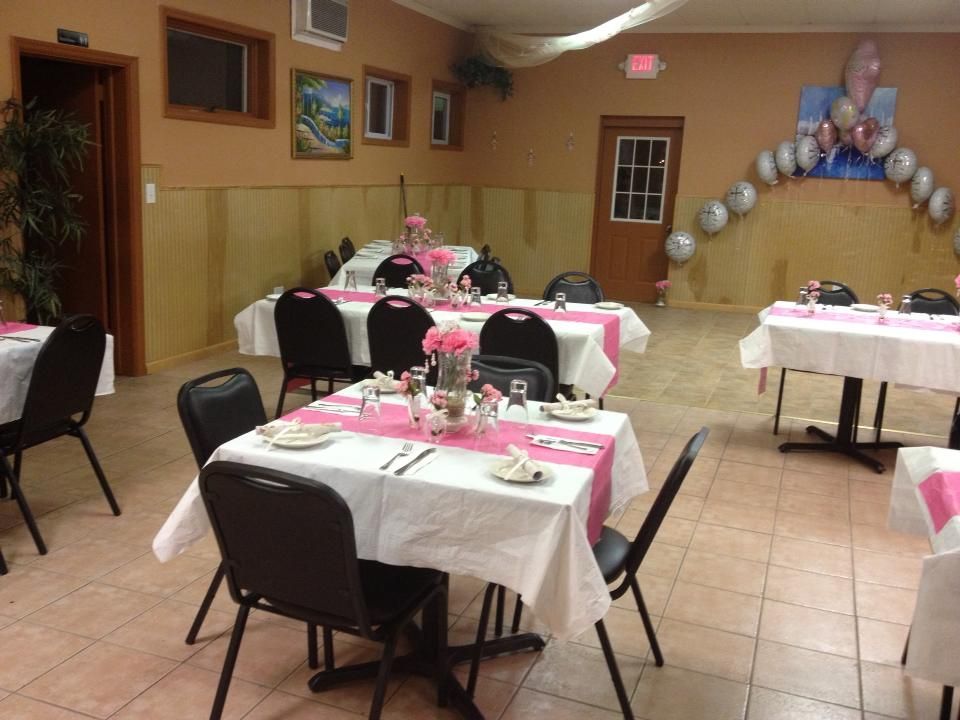 A room decorated for a party. Tables with white tablecloths and pink runners, black chairs, and balloon arch.
