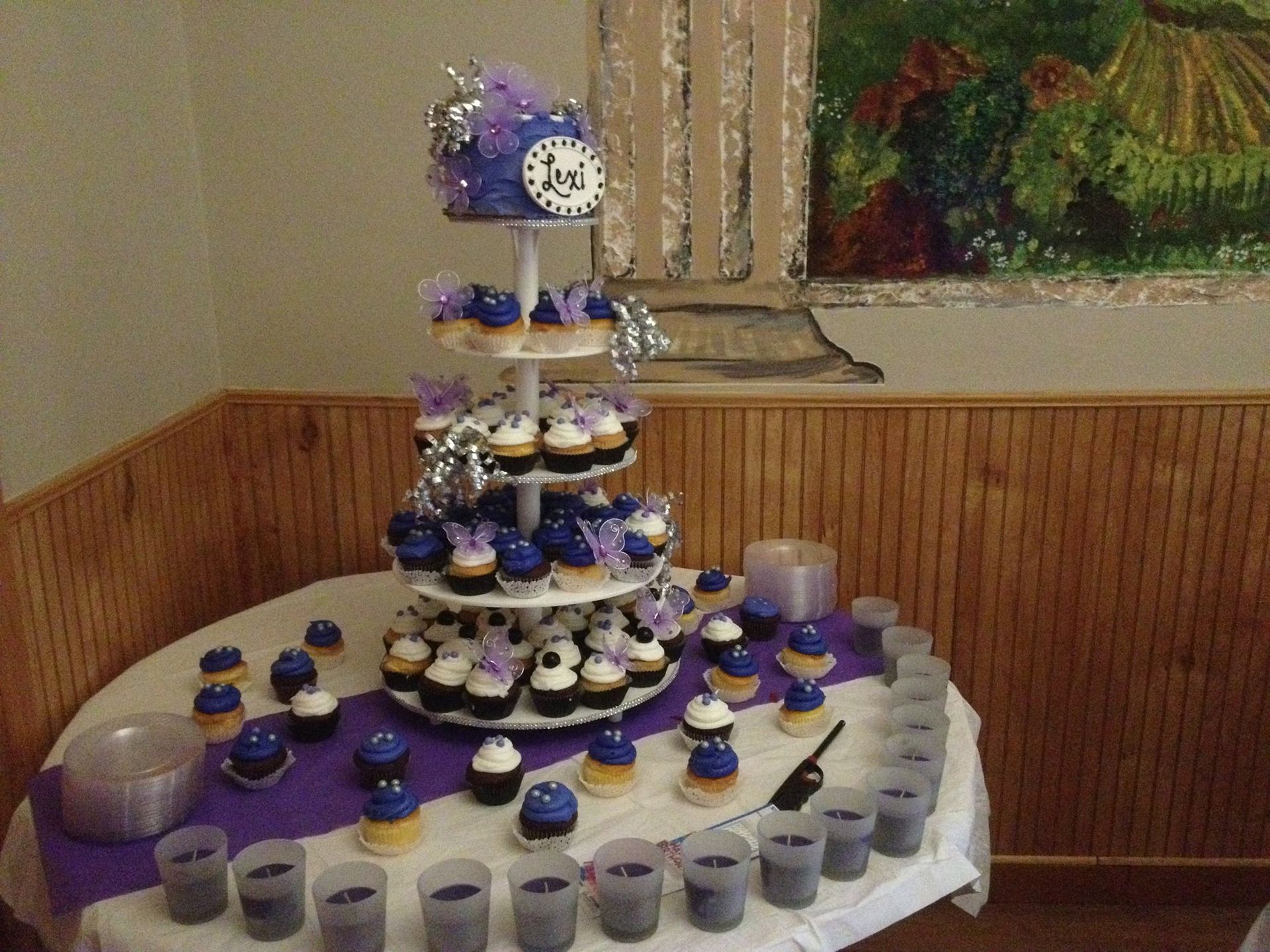 A tiered cupcake stand with purple and blue frosted cupcakes on a table with small drinks, in a wood-paneled room.