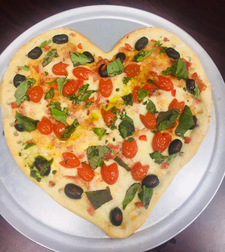Heart-shaped pizza on a metal tray, topped with tomatoes, olives, and basil.