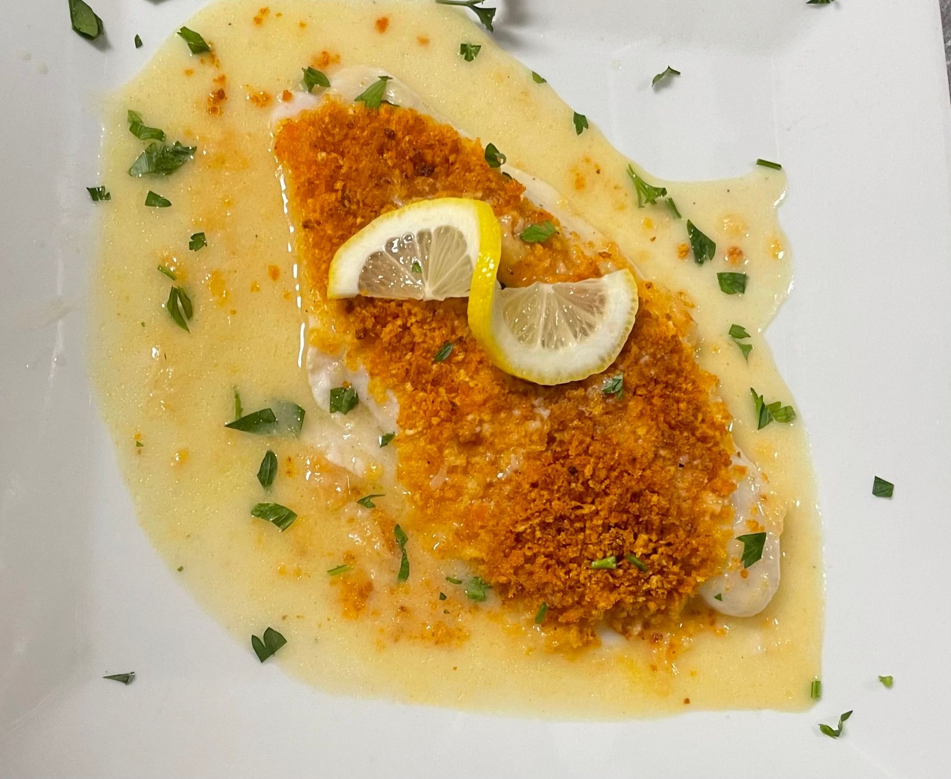 Fish fillet with breadcrumb topping, lemon slices, and sauce, garnished with parsley on a white plate.