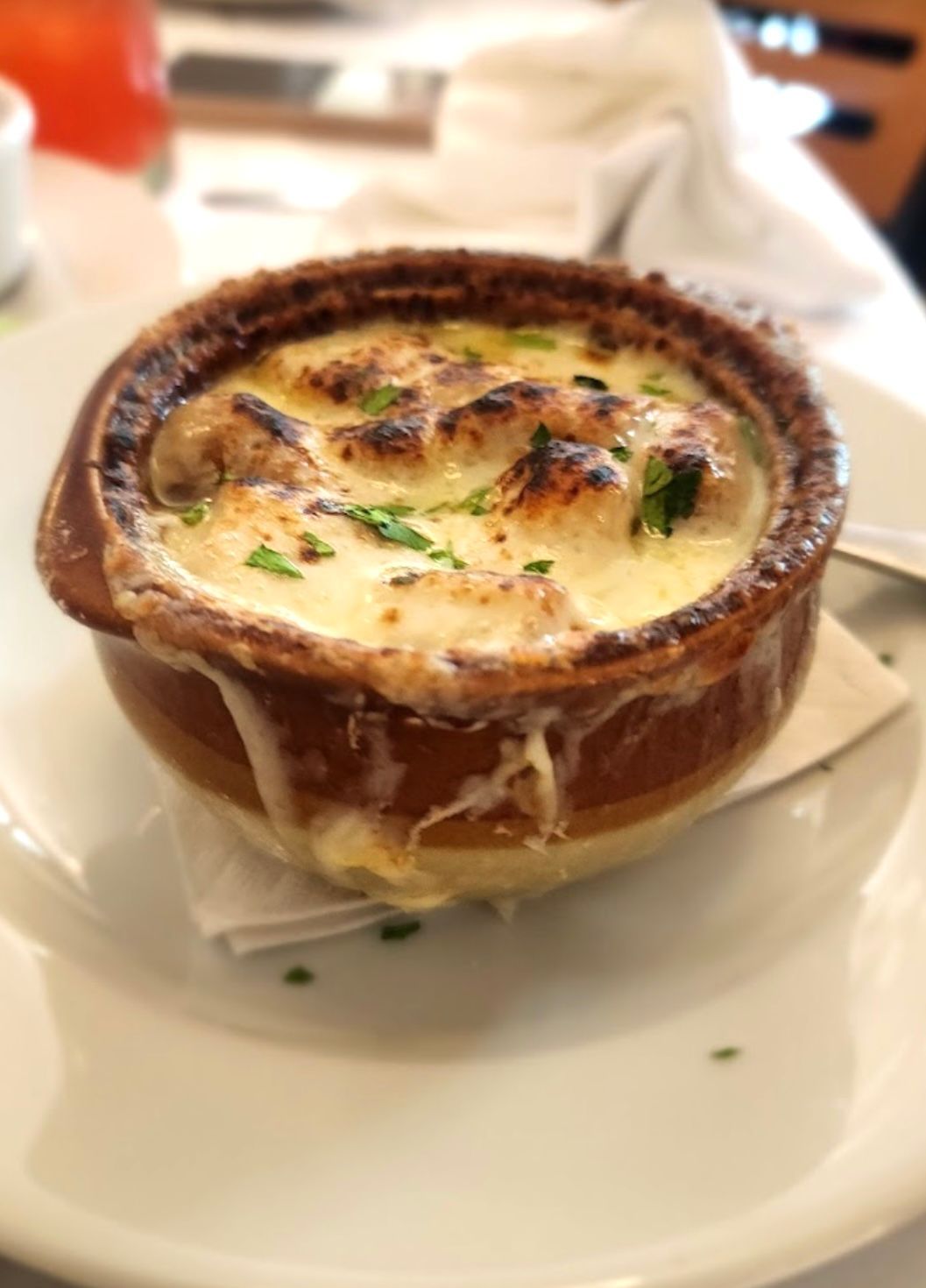Baked French onion soup in a brown ceramic bowl, topped with melted cheese and herbs, on a white plate.
