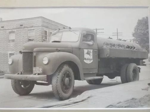 Vintage black-and-white photo of an old flatbed truck parked by a brick building on a street