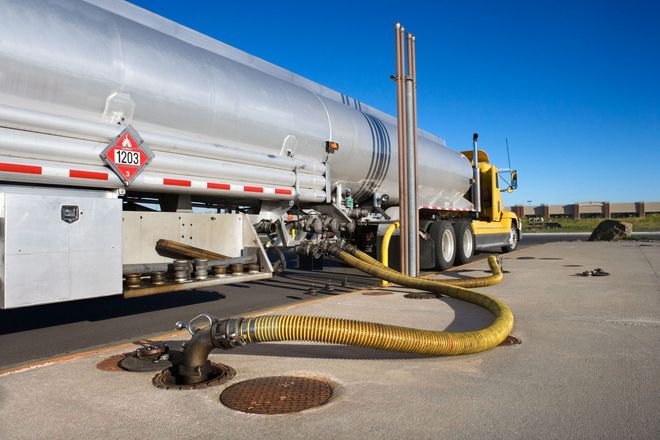 Silver tanker truck with yellow cab parked on pavement, hose connected to a ground fuel pump.
