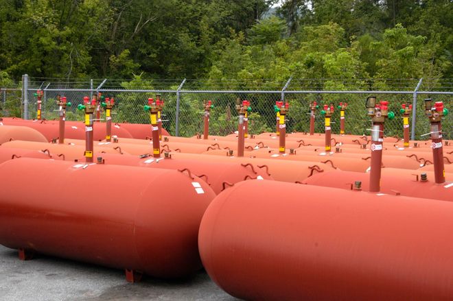 Rows of orange cylindrical tanks with valves, lined up outdoors beside a fence and trees.